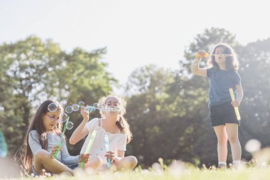 Three beautiful caucasian girls sisters are sitting on a flower lawn in a public park and blowing soap bubbles in the backlight with a blurred background, close-up side view. PARKS REC concept, happy