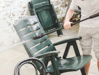 A white guy in gray sweatpants and a T-shirt washes plastic dark green sun loungers under a Karcher faucet in the backyard of his house, close-up side view. The concept of washing sun loungers, house cleaning services.