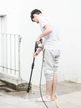 A caucasian guy in gray sports and a T-shirt from the back washes the stone tiles of the terrace in the backyard of his house under the pressure of water from a Karcher, side view, close-up. The concept of washing sun loungers, cleaning services, at 