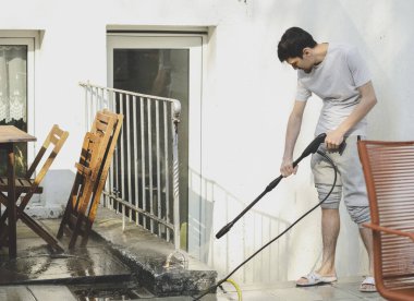 A caucasian guy in a gray T-shirt and sweatpants, standing sideways, washes a curb with a metal fence with a pressure of water from a karcher in the backyard of his house with a karcher, bottom view, close-up. The concept of washing sun loungers, cle