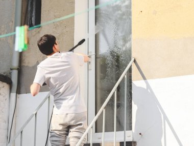 A caucasian guy in gray sports and a T-shirt from the back washes a window with a karcher with water under pressure while standing on a metal staircase in the backyard of his house, side view close-up.The concept of washing the yard,holiday preparati