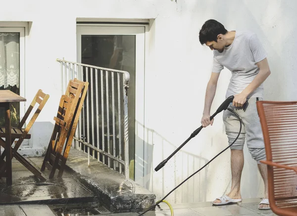 A caucasian guy in a gray T-shirt and sweatpants, standing sideways, washes a curb with a metal fence with a pressure of water from a karcher in the backyard of his house with a karcher, bottom view, close-up. The concept of washing sun loungers, cle