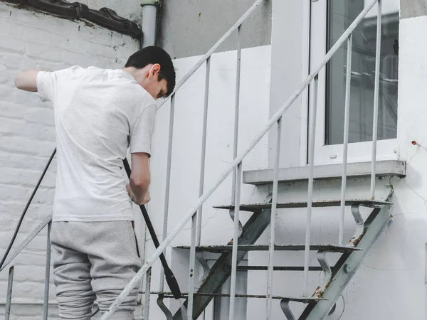 Caucasian guy in gray sportswear and a t-shirt on the side washes with a karcher with water under pressure the steps of a metal ladder up in the backyard of his house, side view close-up.The concept of washing the yard,holiday preparation,at home,cle