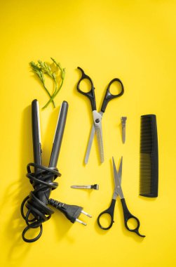 Scissors, combs and a mini hair straightener with green flowers lie on a yellow background, flat close-up. Hairdressing concept.