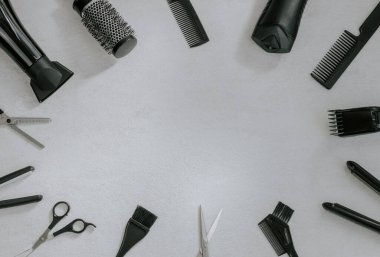 Scissors, hair dryer, hair straightener, combs, brushes, shampoo and hair clipper lie in an oval frame on a gray cement background with copy space in the center, flat closeup. Hairdressing concept.