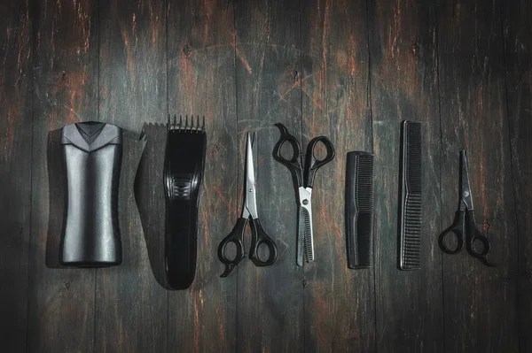 Scissors, combs, shampoo and a men\'s hair clipper lie in a row on a black wooden background, flat lay close-up. The concept of a male barbershop.