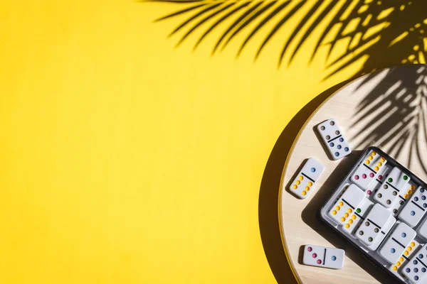 White dominoes with colorful dots in a metal box lie on a round wooden spinning board on the right on a yellow background with a shadow of a palm branch and copy space on the left, flat lay close-up. Concept summer board game.