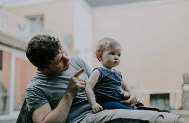 Portrait of caucasian dad with curly brown hair in work uniform pointing with finger talking to a little girl in her arms, looking to the side with an attentive look, sitting on a garden plastic chair in the backyard of his house, close-up side view.