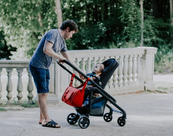 Caucasian young and handsome male dad pulls his hand to a little daughter sitting in a stroller and gently looking at him in a public park on the lawn near the lake, close-up side view. Concept of fatherhood, dads, family vacation, spring walk, dad c