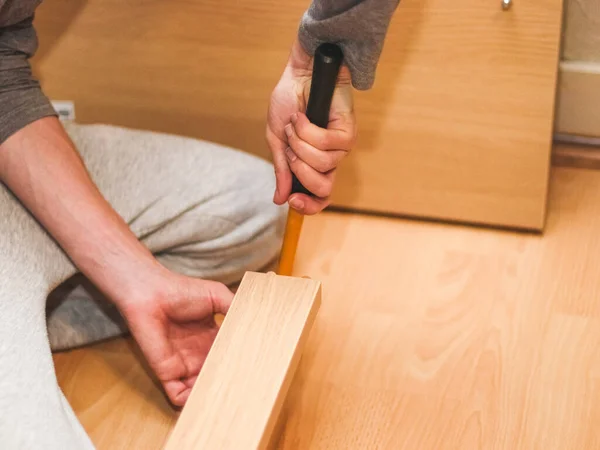 The hands of a Caucasian young man hammer a wooden nail into the headboard with a yellow hammer, sitting on the floor in his room, close-up side view. Concept of assembling furniture, shopping, at home.
