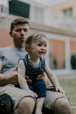 Kafkas bir babanın portresi, iş üniformalı, kıvırcık kahverengi saçlı, kolları bağlı, karamsar ağlayarak yüzünü çeviren, evinin arka bahçesindeki plastik bir sandalyede oturan küçük bir kız.