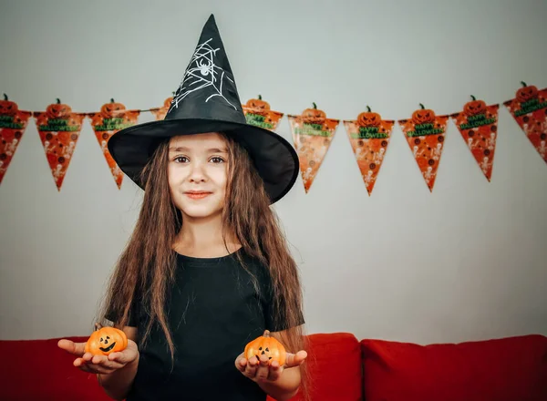 Portrait of a smiling girl in a halloween hat holding two decorative pumpkins in her hands, close-up side view.