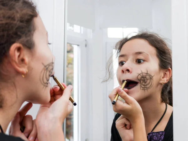 Caucasian teenage girl in a witch dress paints her lips with a black pencil looking in the mirror and preparing for the celebration of halloween,side view close-up.Concept preparation for halloween