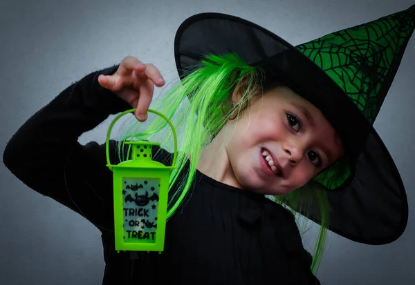 Portrait of a smiling girl in a Halloween hat holding a green lantern in her hands with the inscription: trick of treat on a gray background, close-up side view. Halloween concept, Halloween