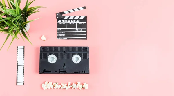 Cinema clapperboard, film strip, videotape, popcorn and flowerpot on the left against a pink background with copy space, top view close-up. Cinema concept.