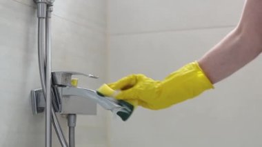 The hands of a caucasian young woman in yellow gloves rub a dirty faucet in the bathroom with a sponge and detergent, side view close-up in slow motion. Cleaning the bathroom concept.