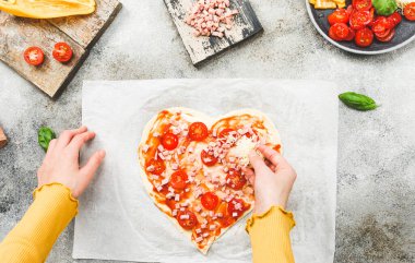 Sevgililer Günü 'nde pizza kalplerine rendelenmiş peynir döken beyaz bir kızın elleri. Masada malzemeler var. Yakın çekim. Sevgililer Günü için pizza pişirme kavramı.