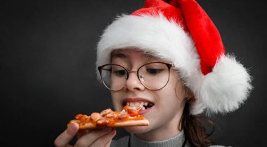 Noel Baba şapkası takan mutlu beyaz kız elinde bir dilim pizza tutuyor ve siyah arka plandaki yazınız için ağzıyla fotokopi aletiyle ısırıyor..