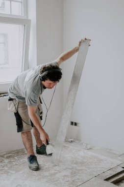 Young handsome caucasian male plasterer with brown curly hair listens to music through headphones and cleans the putty trowel after finishing wall plastering, close-up side view.Concept cleaning trowel, wall puttying, apartment renovation, constructi