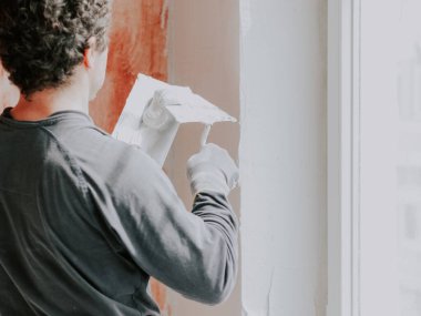 Gri uzun kollu tişört, eldiven ve kıvırcık kahverengi saçlı yakışıklı beyaz bir adam, kırmızı havan topu ve yakın görüş ile hazırlanmış duvarların arka planına karşı bir spatulaya macun koyuyor. Daire ve ev yenileme konsepti, duvar kaplaması