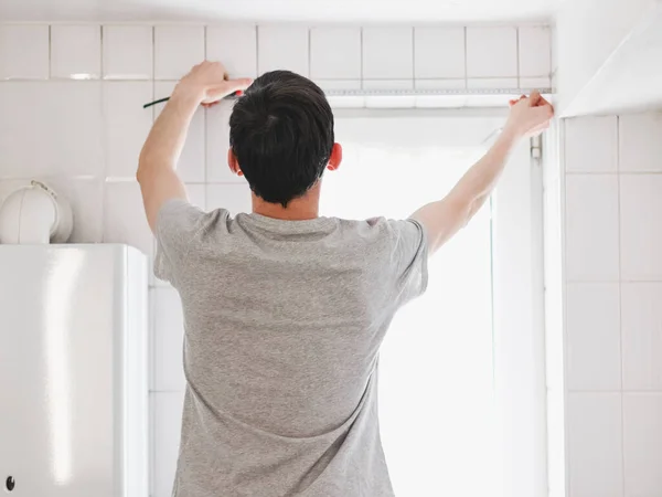A young caucasian guy in a gray t-shirt and brown hair from the back measures the width of a small window in the bathroom with a tape, close-up side view with selective focus. The concept of window measurement, construction work, diy, repair, at home