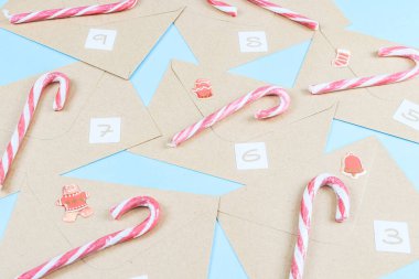 Üzerinde numaralar, şeker lolipopları ve Noel süslemeleri olan Kraft zarfları mavi arka planda, yakın plan yan görüntüde duruyor. Advent takvim kavramı.