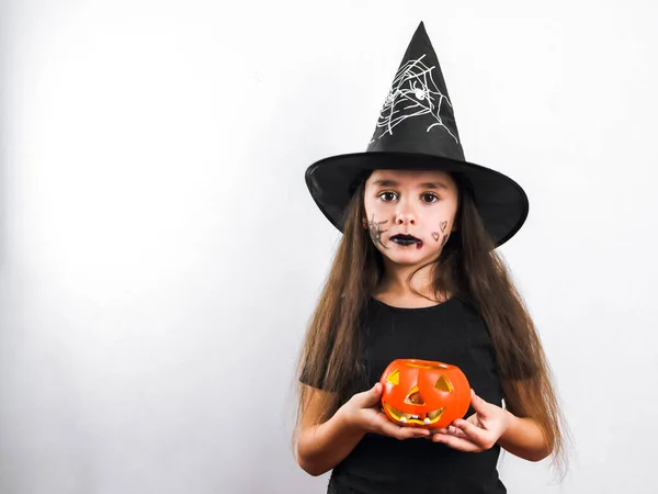 Portrait of a beautiful caucasian brunette girl in black clothes with long hair, halloween makeup and a witch hat holds a ceramic pumpkin with candies in her hands looking at the camera. stands on the right on a white background with copy space on th