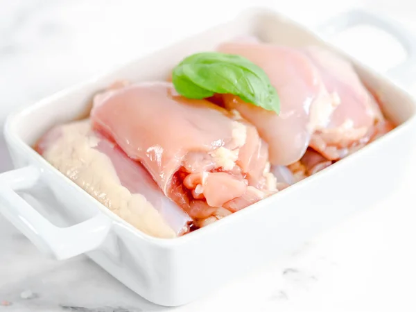 Raw chicken thighs with fresh basil leaf lie in a gray ceramic dish on a marble table, close-up side view.Raw food concept.