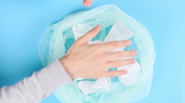 Genç beyaz bir adamın elleri beyaz plastik bardakları sıkar ve mavi arka planda plastik bir torbaya koyar..