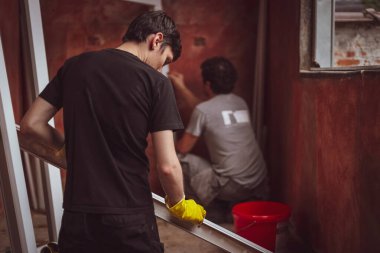 Arkadan, inşaat üniformalı iki beyaz erkek pencere çerçevelerini pencere montajı için hazırlamak üzere kırmızı duvarlı bir balkonda yıkıyor. Ev onarımı, pencere kurulumu, yorumlama