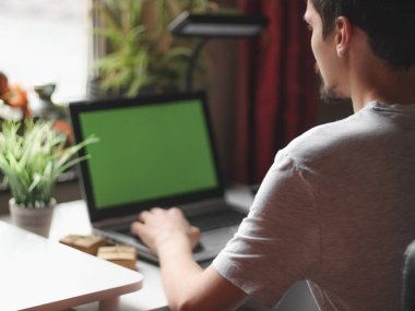 Gri tişörtlü genç beyaz bir adam online alışveriş yapıyor, odasının arkasındaki sandalyesinde oturuyor penceresinin önündeki dizüstü bilgisayarlı yeşil ekranlı, saha derinliğinde yan görüntüsü olan. Evde online alışveriş kavramı.,