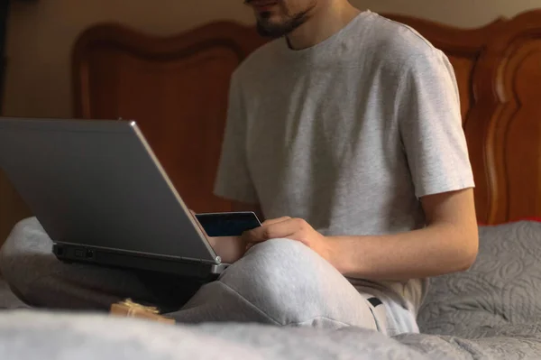 A young caucasian man in gray sweatpants and a t-shirt holds a bank card in his hand and type on the keyboard of a laptop that he holds on his knees, and next to it is a small kraft gift box, sitting on the bed in the bedroom, close-up side view with