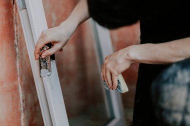 Kafkasyalı genç adam, çamurdan bir pencere çerçevesini inşaat bıçağıyla temizliyor, elinde sabunlu, kirli bir sünger tutuyor, seçici bir odak ile yakından bakıyor. Ev yenileme konsepti, pencere çerçeveleri.