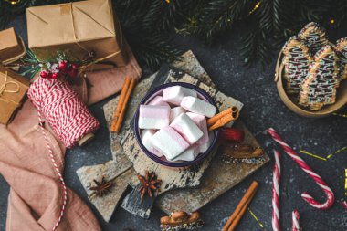 Sıcak kakaolu bir kırmızı demir kupa, tarçınlı marşmelovlar, kesme tahtasında zencefilli adam ve köknar dalları olan koyu çimento arka planda süslemeli Noel şekeri, yakın çekim. Tatil içkisi konsepti.