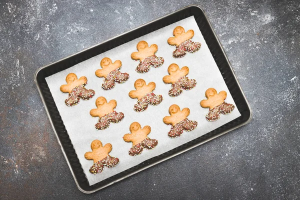 Three rows of gingerbread men in a baking dish with baking paper lie in the middle against a dark cement background, top view close-up. Holiday food concept.