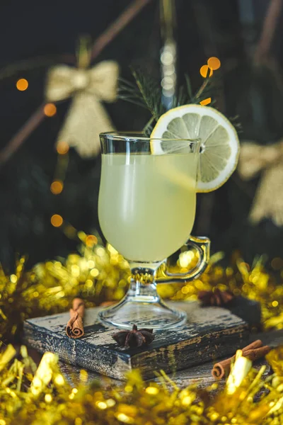 A glass of lemon drink with a circle of lemon and cinnamon sticks on a cutting board with golden tinsel around on a blurred holiday background, close-up side view. Holiday drink concept.