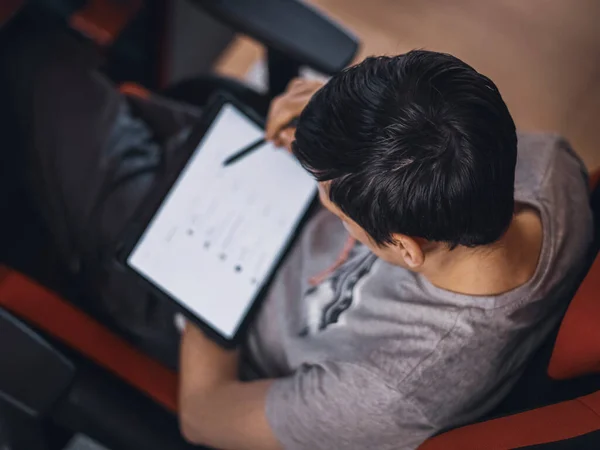 A young caucasian guy in a gray t-shirt sits in a work chair and browses an online menu for ordering takeaway food on a tablet in a blurry, top view close-up. Concept using technology, social media, online chatting, modern lifestyle, at home, online