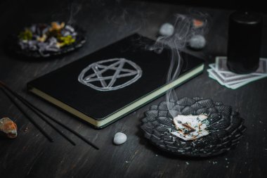 Bir tutam dumanlı siyah metal bir fincan tabağı, sihirli beyaz sembollü bir deri kitap, siyah mumlar, tarot kartları, kurumuş çiçekler ve taşlar siyah ahşap bir arka planda, yakın plan görüşünde. Esrarengiz kavram, karanlık tarz..