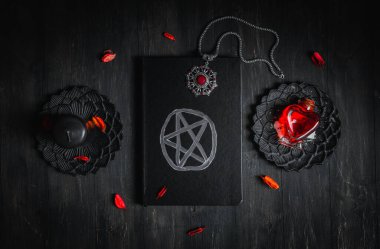 Sihirli beyaz sembollü siyah deri bir kitap, siyah bir mum, kırmızı taşlı bir tılsım, kurumuş bir çiçek ve metal fincan tabağı şeklinde bir kalp şeklindeki kırmızı zehir siyah ahşap arka planda yatar, düz bir yakın plan. Esrarengiz bir kavram.