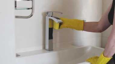 Sarı eldivenli beyaz bir kadının elleri uzun bir lavabo musluğunu deterjanla yıkıyor, yan görüntüyü yavaş çekimde çekiyor. Banyoyu temizleme fikri..