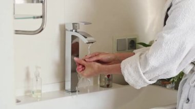 Beyaz bornozlu beyaz bir kadın ellerini temiz su deresinin altında duruluyor ve banyodaki musluğu parmaklarıyla kapatıyor, yavaş çekimde yandan bakıyor. Vücut bakımını kabul et