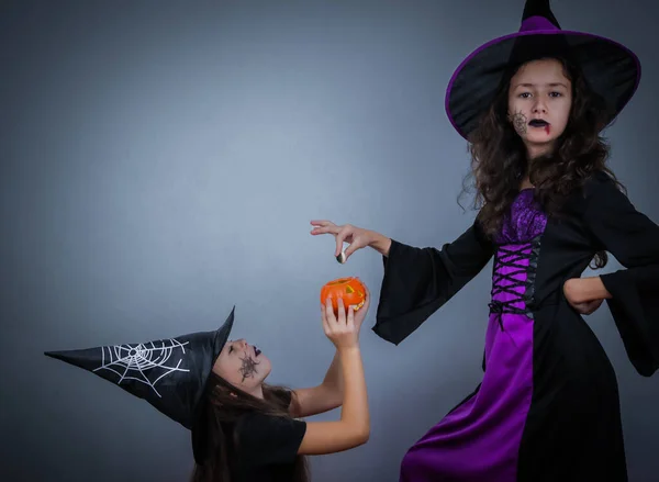 Two beautiful caucasian brunette girls in witch costumes with long hair, halloween makeup in witch hats hold a ceramic pumpkin in their hands and share jelly candies looking at the camera on a dark