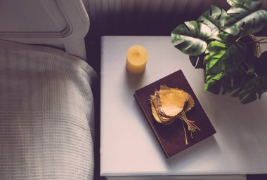 Kahverengi kitap, sonbahar kavak yaprakları ve yatağın yanındaki beyaz komodinin üzerinde sarı bir mum, tarlanın derinliği ile yakın plan. Sonbahar tatili kavramı.