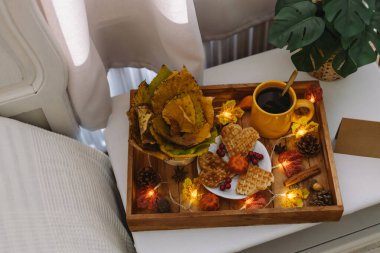 İçinde bir kupa kahve olan ahşap bir tepsi, tabakta waffle, sepette kavak yaprakları, sonbahar süsü ve pencerenin yanındaki beyaz bir komodinin üzerinde yanan bir çelenk, tarlanın derinliği ile yakın çekim. Sonbahar tatili ve kahvaltı konsepti.