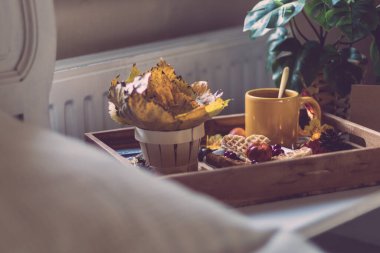 Tahta tepside bir kupa kahve, tabakta waffle, sepette kavak yaprakları, tencerede bir manster ve beyaz bir komodinin üzerinde yanan bir çelenk, tarlanın derinliği ile yakın çekim. Sonbahar tatili ve kahvaltı konsepti.