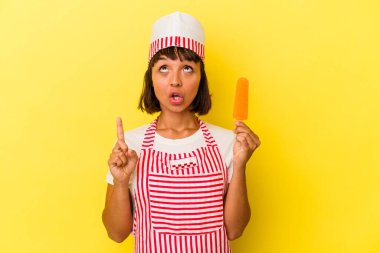 Genç melez dondurma üreticisi elinde dondurma tutan kadın sarı arka planda izole edilmiş ve ağzı açık bir şekilde yukarı bakıyor..