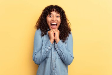 Genç melez kadın sarı arka planda izole edilmiş şans için dua ediyor, şaşırmış ve ağzı açık bir şekilde önüne bakıyor..