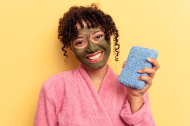 Sarı arka planda izole edilmiş duş süngeri tutan pembe bornoz giyen genç bir ırk kadını mutlu, güler yüzlü ve neşeli..