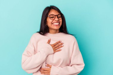 Mavi arka planda yalnız yaşayan genç Latin kadın mutlu bir şekilde gülüyor ve ellerini karnının üstünde tutarak eğleniyor..