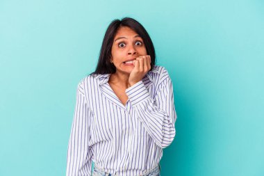 Mavi arka planda izole edilmiş, tırnak yiyen, gergin ve endişeli genç bir Latin kadın..